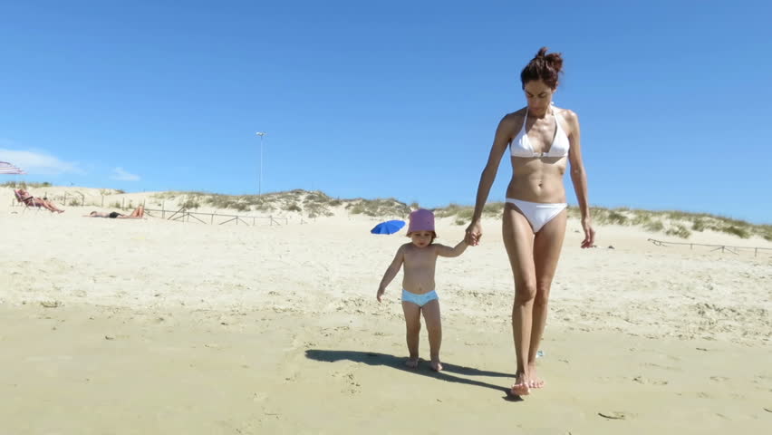 two years age baby with red hat and blue swimwear with brown hair mother in white bikini walking on sand at beach
