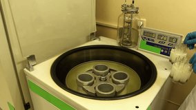 A man in white clothing and blue gloves loads glassware into centrifuge, starts, stops and partly unloads this device.  Opened transparent centrifuge cover. No camera movement.