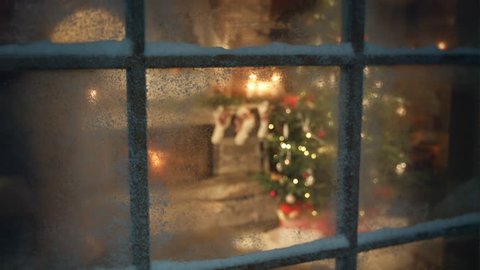 Christmas tree and fireplace scene through frozen and snowy window dolly shot. Decorated christmas tree in cozy and warm farm house and presents under the tree while snowing outside.