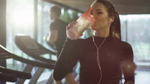 Attractive caucasian girl is drinking a protein shake drink next to a treadmill in the sport gym. Shot on RED Cinema Camera in 4K (UHD).