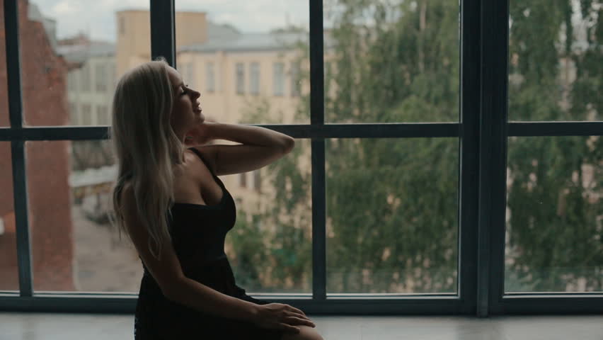 Attractive woman in dress sitting on a wide windowsill and looking in the window