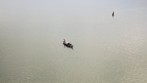 Upper distance view on a small fishing boat on river and fisherman lifting his net out of water; Other fishing boats around proceeding down the river 