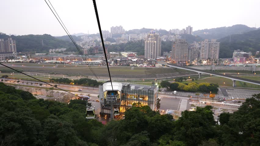 Cablecar cabin approach to lower terminal station, cityscape on background, aerial view. Maokong gondola lift system, transportation to Maokong area from MRT station Taipei Zoo, Taipei city suburb
