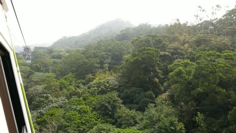 Rainforest at hilly landscape, view from moving Maokong gondola lift. Cloudy day, foggy view of foresty mountain landscape. Cable car travel above the wild nature, pass tree crown.