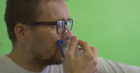 Man is Glasses on Chroma Key, Green Screen, Patient in White T-Shirt is Keeping a Nebulizer Mask at His Face, Breathing through Inhaler, Steam from Inhaler, Man with Asthma, Sad Serious Face and