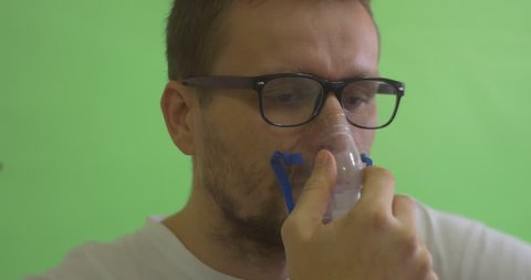 Man is Glasses on Chroma Key, Green Screen, Patient in White T-Shirt Has Put a Nebulizer Mask to His Face, Breathing through Inhaler, Man with Asthma, Serious Face, seriously sick man, breathing