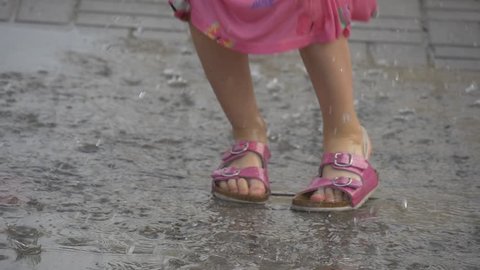 Close-up on camera filmed the feet of a little girl. She's having fun and jumping through the pudle. A sandals have completely got wet. The asphalt is wet after a rain.