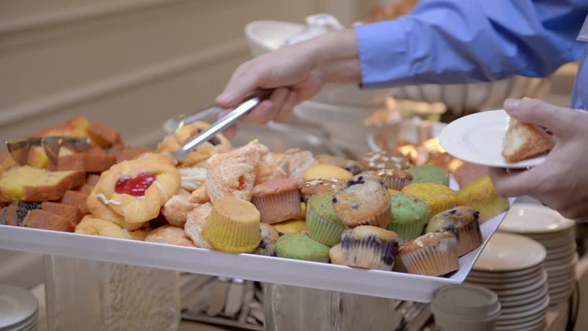 A guest attending a corporate business seminar in a hotel helps themselves to the free breakfast of cakes and sweets at the catered buffet table.