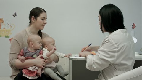 Female pediatrician doctor's explaining something to mother with newborn twins and writing a prescription for drugs