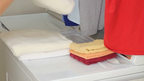 Arms and hands of a male come in from the left wearing light clothes, folding a towel onto two other towels on top of a dryer in a laundry room. Then he picks up the towels and exits to the left. 