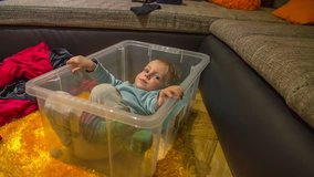 In this video, we can see that a cute little boy is tired of playing in a plastic box and he slowly gets up in order to get out of the box. Wide-angle shot.