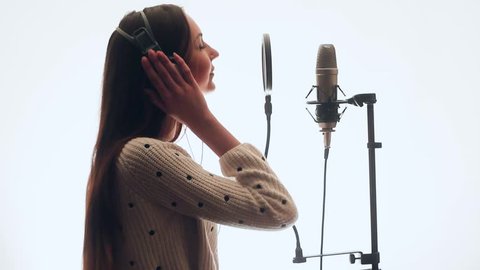 Sweet singer sings into a microphone in a recording studio. She hops. The girl is very beautiful. It stands on a white background. Backlight.
