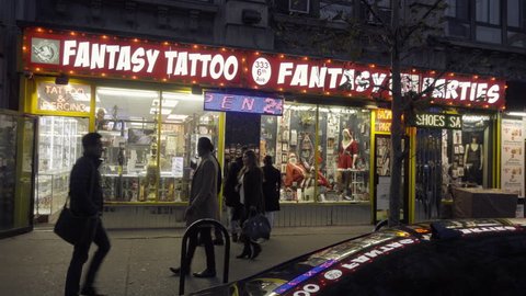 NEW YORK - DEC 6, 2015: famous Fantasy Tattoo party store, neon sign, storefront, West Village, silhouettes New Yorkers walking, night NYC 4K. Greenwich Village is a famous neighborhood in the city.