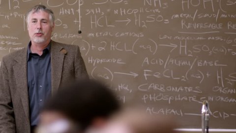 A professor pointing to chemistry equations on a chalkboard, with students in the foreground Stock Video