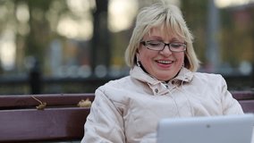 Blonde woman with Tablet PC sits on the bench and talks on video chat. Female with Tablet-PC. Senior woman sits on the bench near the road and communicates via tablet computer. She is in a merry mood