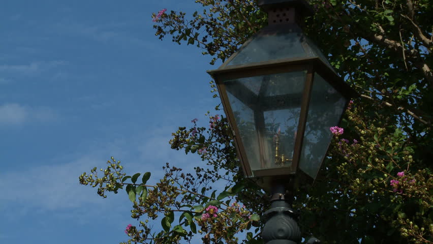 A gas light fire flame street lamp in Vicksburg, Mississippi. Pretty pink flowers bloom on the tree with blue sky background. Southern small town Americana.