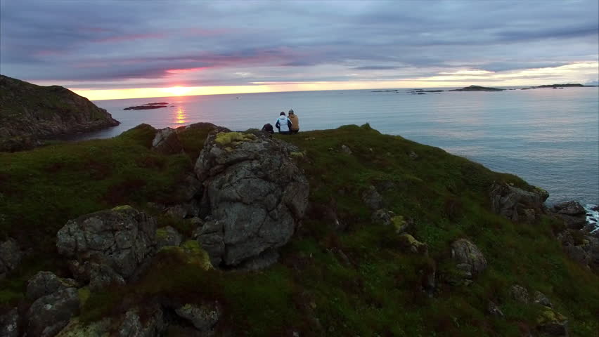 Young couple sitting by the ocean and watching midnight sun on Vesteralen islands in Norway. Aerial 4k Ultra HD. Royalty-Free Stock Footage #13710527