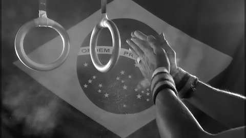 Taped hands of gymnast clapping white chalk powder into a cloud under gymnastic rings in front of a Brazil flag background in gritty black and white