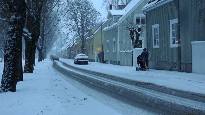 Heavy snowfall on street in old suburb. Cars driving slowly.
Woman with baby carriage passing.
