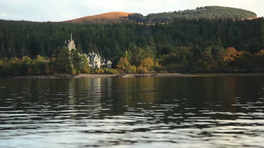 A beautiful stone castle sits at the foot of a rolling green hill overlooking a lake.