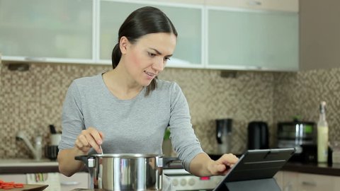 Young woman cooking with help of recipe on tablet and testing the food in the kitchen