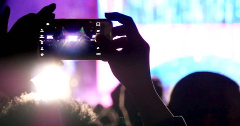 Fan person taking video and photos on mobile smart phone at concert party crowd cheering at rock music event with flashing light show and band on stage.