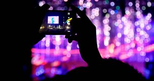 Fan person taking video and photos on mobile smart phone at concert party crowd cheering at rock music event with flashing light show and band on stage.