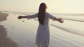 Rear view video of woman standing with arms outstretched on shore. Tracking slow motion shot of carefree female with eyes closed. Woman in white sundress enjoying nature on beach during sunset.
