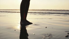 Low section footage of woman walking on wet sand. Slow motion video of female with bare feet on shore during sunset. Tracking shot of woman strolling. Sunlight is reflection on seascape.