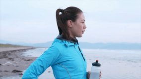Slow motion video of fit woman enjoying fresh air on beach. Sporty female is in blue jacket. She is looking away at breaking waves while drinking water on shore.