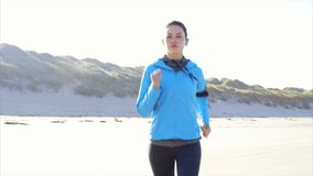 Slow motion video of confident woman running on beach. Determined fit young female is listening music while jogging on sunny day. Beautiful female is exercising in nature.