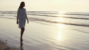 Slow motion video of woman walking barefoot on sea shore. Tracking shot of female enjoying nature during sunset. Full length of female in white sundress. Reflection of sunlight is falling on seascape.
