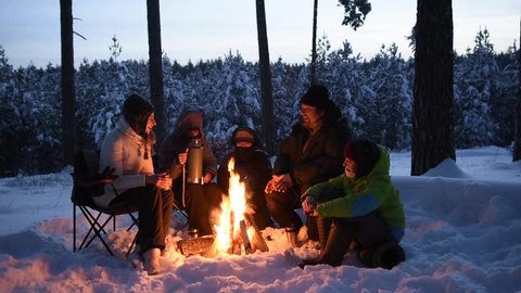 Sitting, laughing and drinking tea  around a campfire with family and friends in winter forest