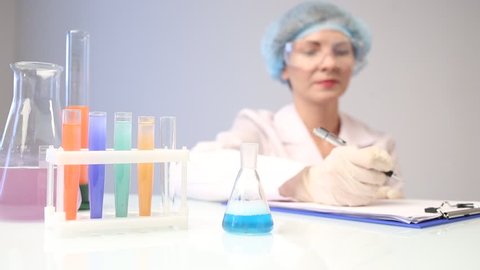 female chemist pouring a blue liquid into a chemical flask. writes the results