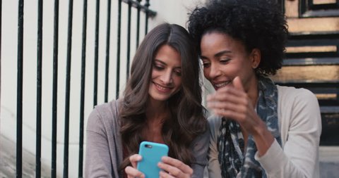 Two beautiful woman friends sitting on steps having fun talking selfie on smart phone happy friendship lifestyle