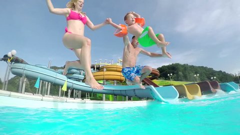 SLOW MOTION CLOSE UP: Happy excited young family running and jumping into the pool water in aqua park on a beautiful summer day on their holidays