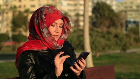 Arabic muslim woman wearing red scarf, sitting on a park bench and using mobile smart phone