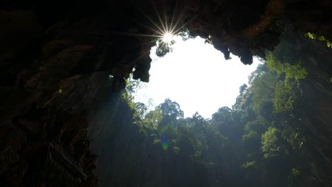 Impressive sunshine through open cave ceiling, blazing sunstar against black frame of cave vaults, green trees on gap edges, sun shine through foliage and leaves while camera panning move left.