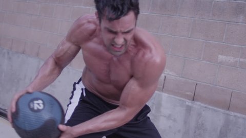 A strong, ripped, cut, buff man doing medicine ball exercises in slow motion, bouncing the ball off of a wall outside - fitness / crossfit / exercise / workout