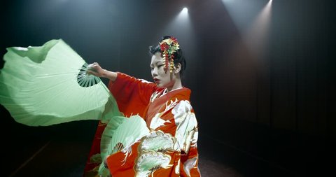 beautiful Japanese geisha posing for the camera with fans on stage, epic slow motion, smoke and dark background