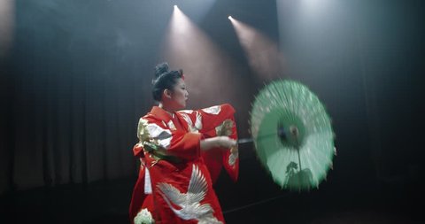 beautiful Japanese geisha posing for the camera with an umbrella on stage, epic slow motion, smoke and dark background