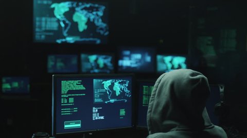 Male hacker in a hood works on a computer with maps and data on display screens in a dark office room. Shot on RED Cinema Camera in 4K (UHD).
