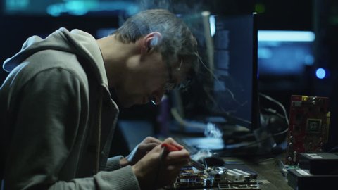 Electronics engineer is soldering an electric board with processors in a dark office with display screens. Shot on RED Cinema Camera in 4K (UHD).