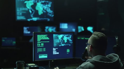 Male hacker in a hood works on a computer with maps and data on display screens in a dark office room. Shot on RED Cinema Camera in 4K (UHD).