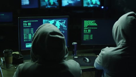 Two hackers in hoods work on a computers with maps and data on display screens in a dark office room. Shot on RED Cinema Camera in 4K (UHD).