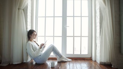 Young woman at home sitting on modern chair in front of window relaxing in her living room using tablet pc