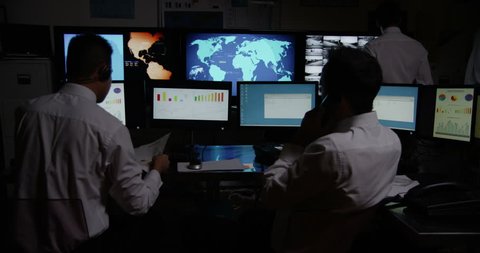 4k / Ultra HD version A team of male security personnel are manning the stations within a busy system control room. It is very dark and the characters are silhouetted against the screens. 
