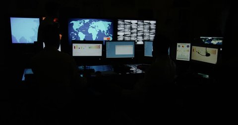 4k / Ultra HD version A team of male security personnel are manning the stations within a busy system control room. It is very dark and the characters are silhouetted against the screens. Slow motion