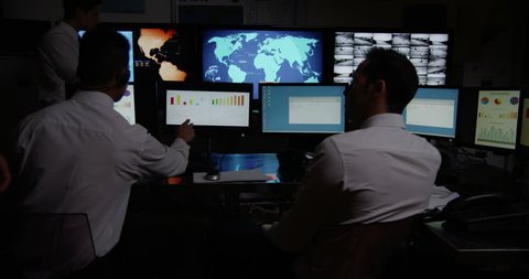 4k / Ultra HD version A team of male security personnel are manning the stations within a busy system control room. It is very dark and the characters are silhouetted against the screens.