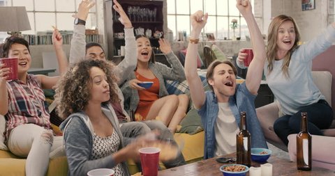 Happy diverse group of student sports fans throwing arms up in excitement celebrating goal watching sports event on TV together bonding as friends eating snacks drinking beer